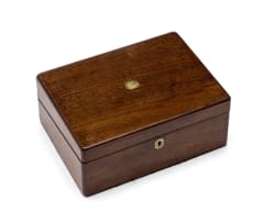 Wooden writing box with a brass inlay in the centre of the lid, brass hinges and small key, opening into two halves onto a red velvet rectangular surface. A latch opens the top half with a shallow storage space below for writing paper, envelopes and stamps. The bottom half consists of two parts, the bottom part a shallow drawer with a removable lid for storing pens, flanked by two cubicle spaces for storing ink bottles, the top part opens onto an